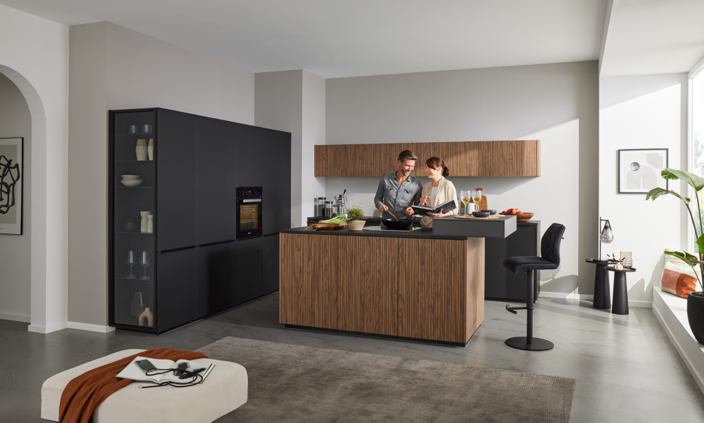 Moderne Küche in Schwarz‑ und Holzoptik mit zentraler Kochinsel, auf der frisches Gemüse und Küchenutensilien liegen. Ein Paar bereitet gemeinsam eine Mahlzeit zu, während Tageslicht durch ein großes Fenster fällt. Die Szene wirkt warm, einladend und zeigt eine hochwertige, zeitgemäße Küchenplanung mit gemütlicher Sitzecke im Hintergrund.