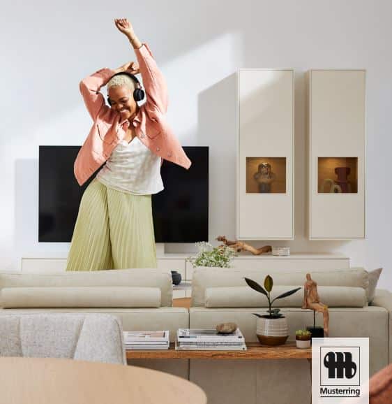 „Person tanzt fröhlich auf einem beigefarbenen Sofa in modernem Wohnzimmer mit Musterring-Möbeln; helle Einrichtung mit TV-Wand, Deko und Tageslicht.