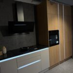 Minimalistische Küche mit schwarzem Induktionskochfeld, modernem Wandhauben‑Dunstabzug, grauen Unterschränken mit LED‑Licht und hohem Holzschrank mit integriertem Backofen.