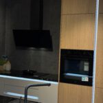 Moderne Ausstellungsküche mit schwarzem Spülbecken, Induktionsfeld, Backofen und hellen Holz‑Weiß‑Fronten, ergänzt durch dekorative Pflanzen und LED‑Lichtakzente.