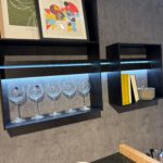 Designorientierte Küchenwand mit zwei schwarzen LED‑Regalen, Weingläsern, Artprints, Büchern und weißen Schalen auf einer schwarzen Granit-Arbeitsplatte.