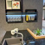 Moderne Küchenwand mit schwarzer Granit-Arbeitsplatte, integriertem LED‑Regal, Weingläsern und zwei gerahmten Artprints; daneben ein Holztablett mit Pflanze und Schalen.