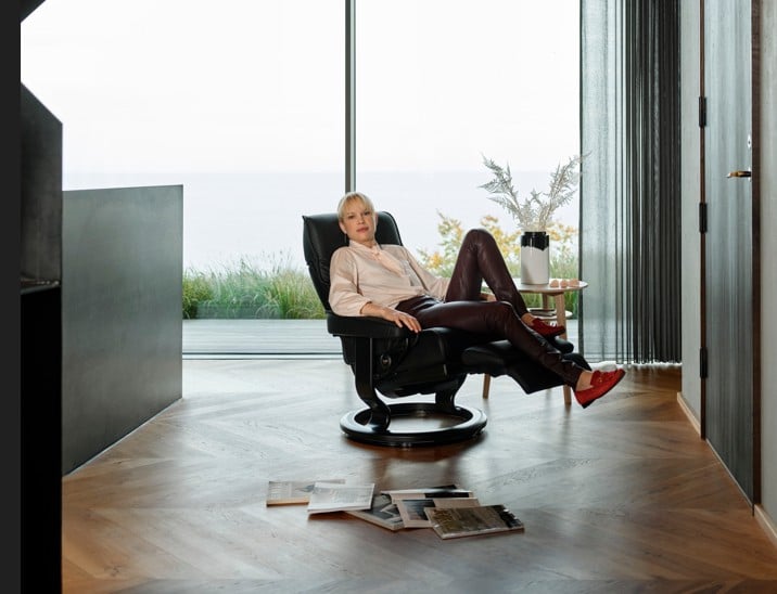 Modernes Wohnzimmer mit bodentiefem Fenster und Blick auf Wasser und Grünflächen. Eine Person sitzt entspannt in einem schwarzen Relaxsessel, umgeben von warmem Holzfußboden, einem kleinen Beistelltisch mit Vase und verstreuten Magazinen auf dem Boden.