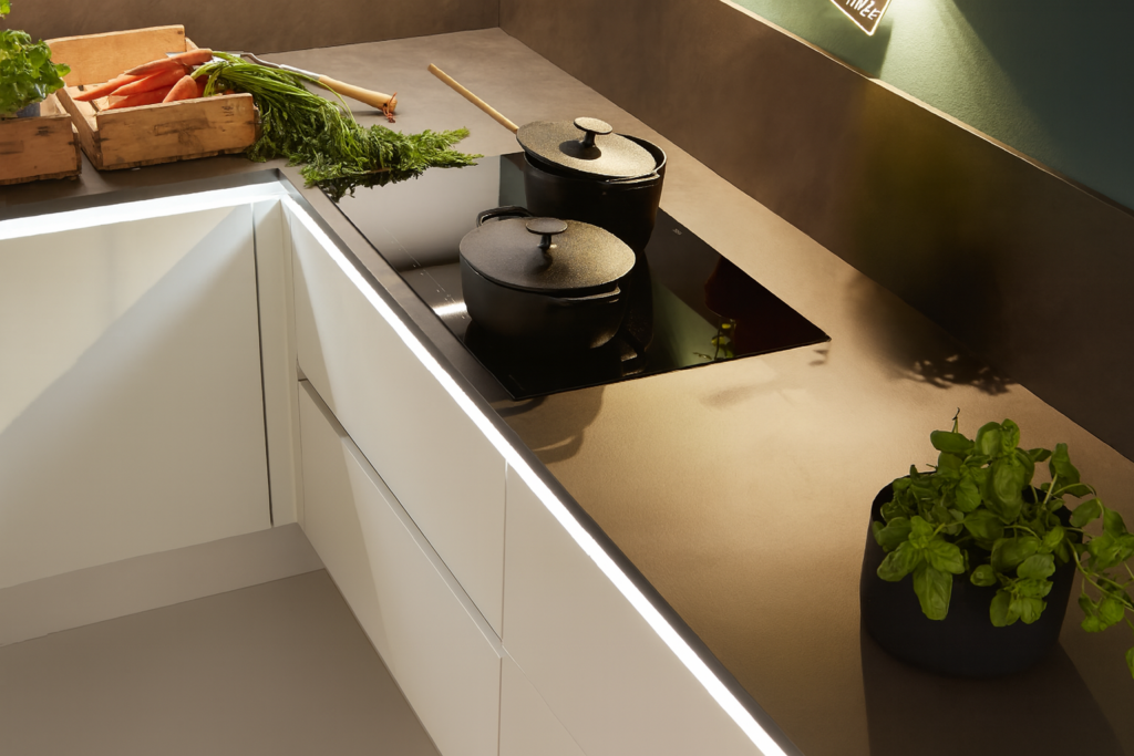 Moderne Küchenarbeitsplatte mit zwei schwarzen Gusseisentöpfen auf einem Induktionskochfeld, frischen Karotten in einer Holzkiste links und einem Basilikumtopf rechts; minimalistisches Design mit weißen Schränken und indirekter Beleuchtung.