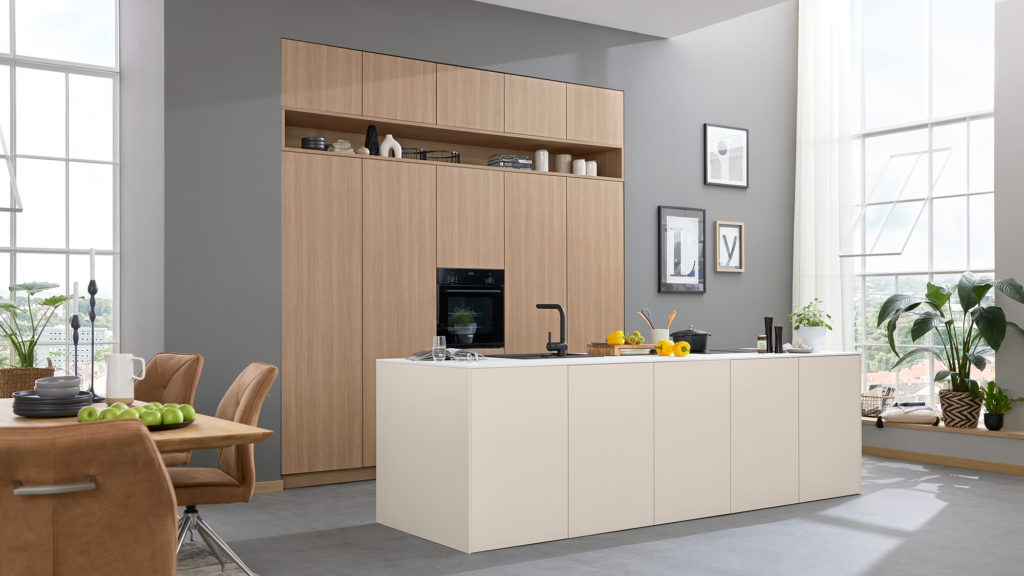 Moderne Küche mit großer weißer Kochinsel, Holzschränken, offenen Regalen und viel natürlichem Licht. Auf der Insel liegen Zitronen, Geschirr und Küchenutensilien. Links steht ein Esstisch mit braunen Stühlen, rechts hängen gerahmte Bilder. Mehrere Pflanzen setzen grüne Akzente.
