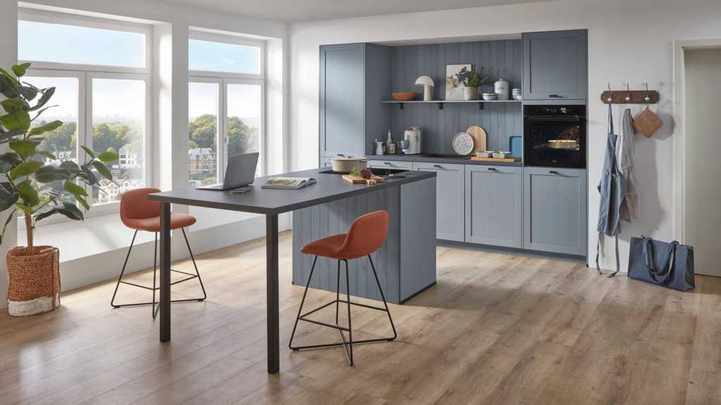 Helle, moderne Küche mit hellblauen Schränken, dunkler Kochinsel und zwei orangefarbenen Barhockern. Auf der Insel liegen Gemüse, ein Topf, ein Laptop und ein offenes Buch. Große Fenster bringen viel Tageslicht hinein, rechts hängen Schürzen an Haken, links steht ein Esstisch mit Stühlen.