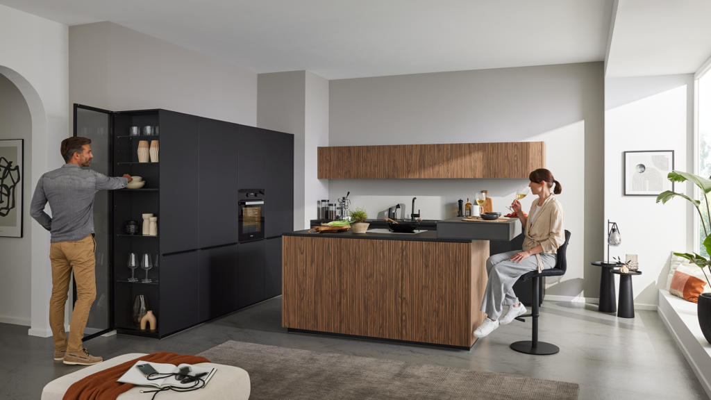 Moderne Küche mit Holz‑Kochinsel und schwarzer Arbeitsplatte. Links öffnet eine Person einen hohen Glasschrank, rechts sitzt eine zweite Person auf einem Barhocker und hält ein Glas Weißwein. Die Küche ist hell, minimalistisch und wird von Tageslicht durch ein großes Fenster erleuchtet.