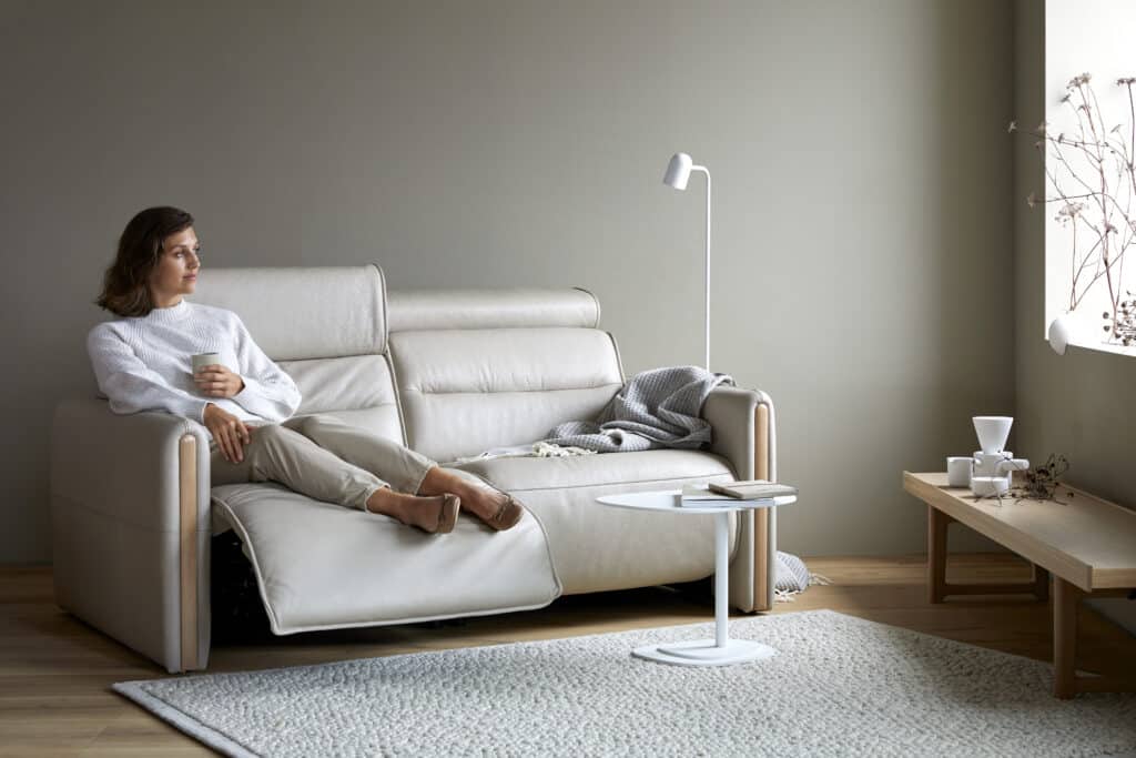 Helles, modern eingerichtetes Wohnzimmer mit beigefarbenem Relaxsessel in Leder, auf dem eine Person mit ausgestreckter Fußstütze sitzt und eine Tasse hält. Daneben ein kleiner runder Beistelltisch mit Buch und Tasse, im Hintergrund eine Holzbank mit Keramikdeko und ein minimalistischer Stehlampe