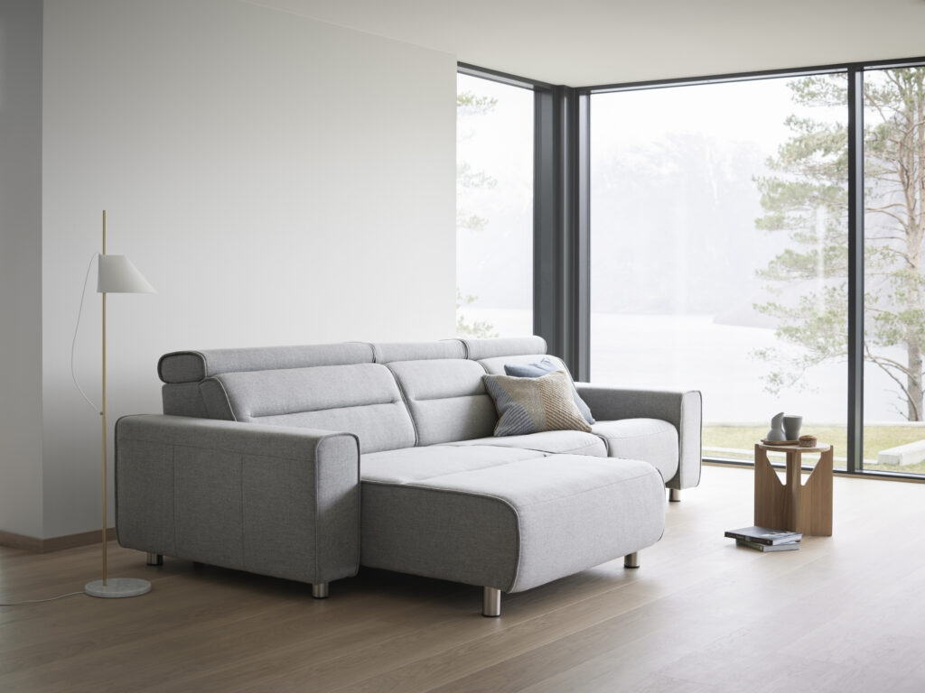 Modernes, helles Wohnzimmer mit einem großen, hellgrauen Ecksofa auf Metallfüßen, dekoriert mit Kissen. Daneben stehen eine schlanke goldfarbene Stehlampe und ein kleiner Holztisch mit Tassen und Tellern. Durch die bodentiefen Fenster fällt der Blick auf eine ruhige Landschaft mit Wasser, Bäumen und Bergen