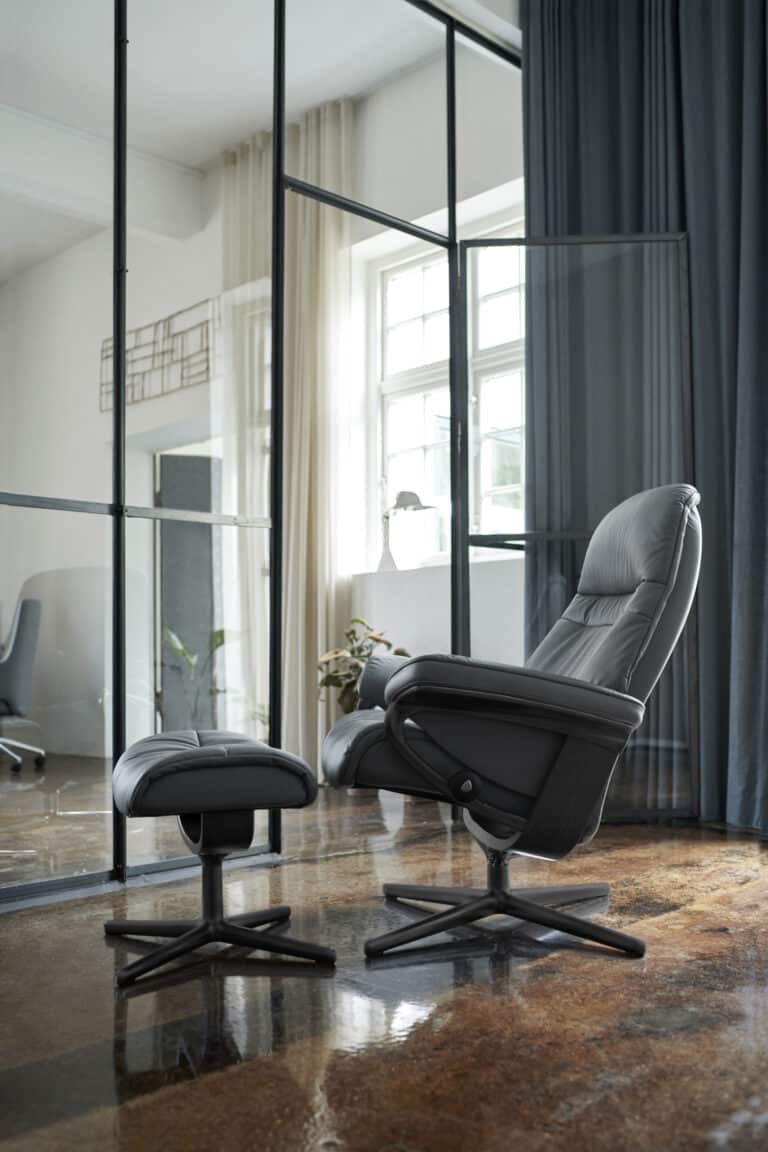 Grauer Stressless Sunrise Relaxsessel mit Hocker vor einem großen Fenster.
