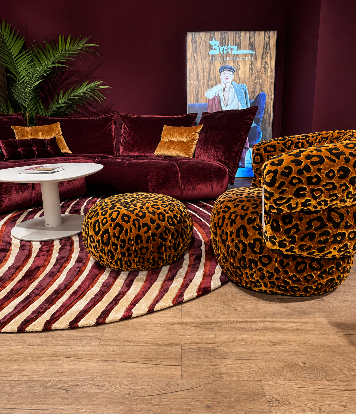 Ein stilvoll eingerichtetes Wohnzimmer mit tiefweinrotem Samtsofa und goldfarbenen Kissen. Davor stehen zwei runde Poufs und ein geschwungener Sessel im Leopardenmuster auf einem gestreiften Teppich in Rot‑ und Cremetönen. Ein weißer runder Couchtisch ergänzt das Ensemble. Im Hintergrund sind eine große grüne Pflanze und ein Poster mit der Aufschrift „Bretz – True Characters“ zu sehen, das eine Person im Vintage‑Look zeigt. Die dunklen Wände und der Holzboden unterstreichen die luxuriöse, warme Atmosphäre.