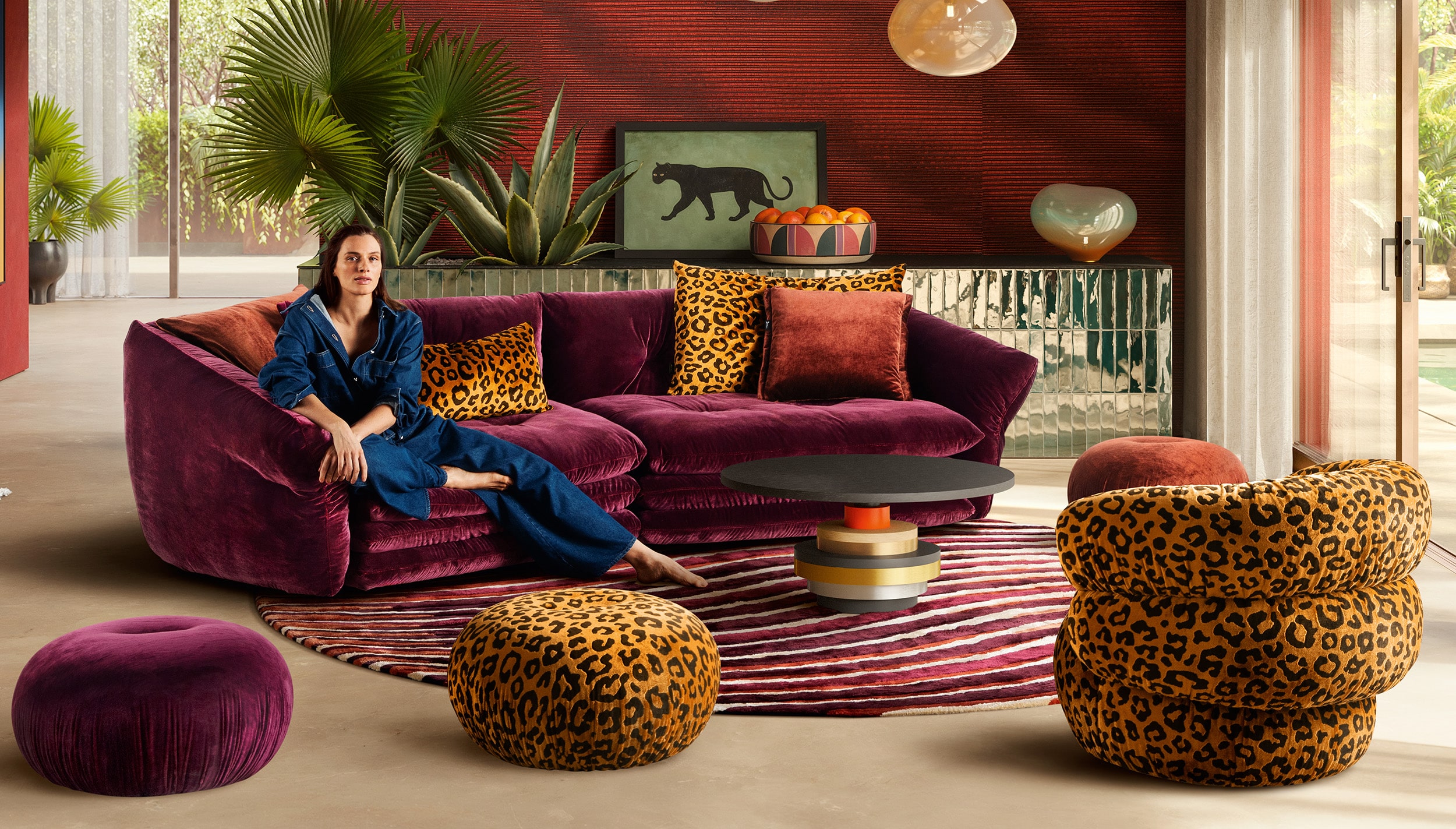 Ein exklusives Wohnzimmer-Setting mit der Bretz Manyara Couch in kräftigem Violett, kombiniert mit Leoparden-Muster-Kissen und Hockern sowie einem runden Teppich im Möbel Homann Designstudio.