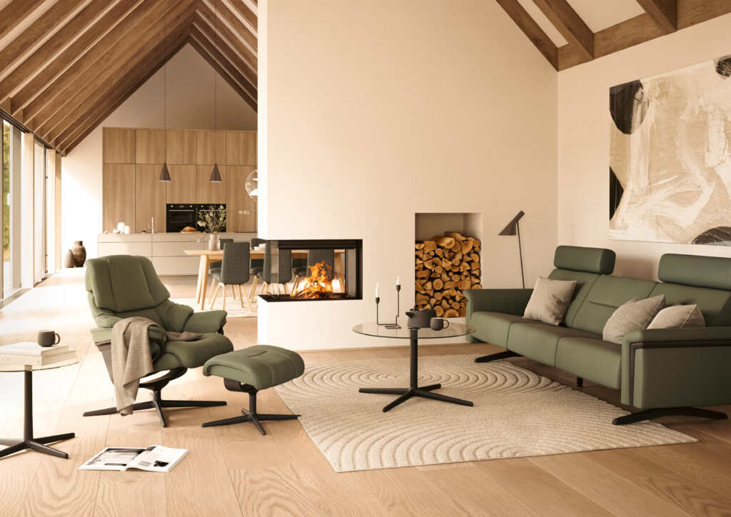 Modernes Wohnzimmer mit grünem Stressless Sofa, passendem Sessel und Hocker, Glas‑Couchtisch, Kaminwand mit sichtbarem Feuer und offener Küche im Hintergrund.