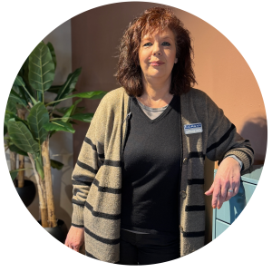 Einrichtungsfachberaterin Claudia Arning steht neben einem hellblauen Möbelstück im Showroom von Möbel Homann, trägt eine beige Strickjacke mit schwarzen Streifen und ein Namensschild. Im Hintergrund eine große Grünpflanze und eine warm getönte Wand.