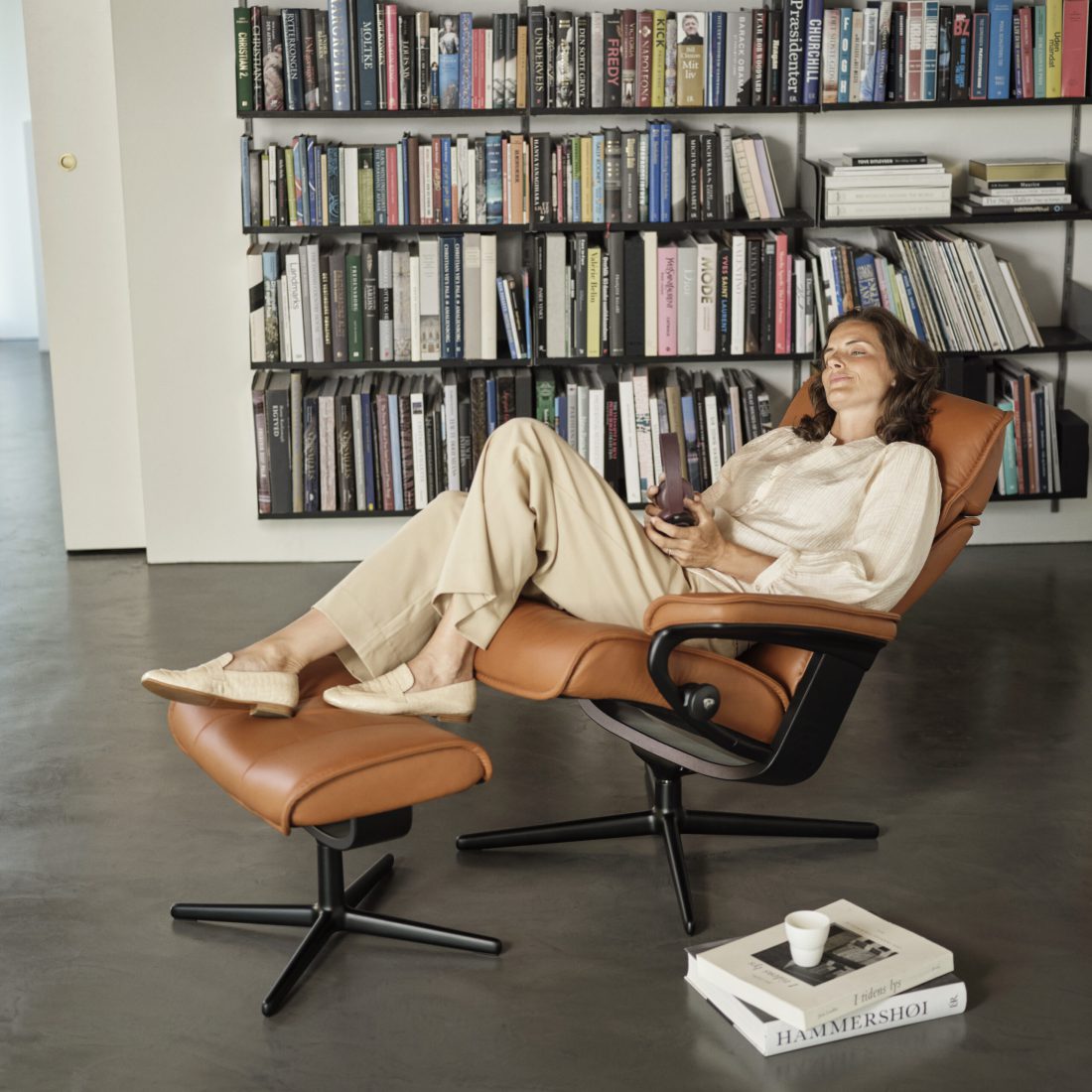 "Ein stilvolles Wohnzimmer mit einem beigen Relaxsessel und einem passenden Fußhocker, beide mit Holzgestell und heller Polsterung. Daneben ein eleganter Beistelltisch mit einer goldfarbenen Oberfläche, darauf einige Bücher und eine dekorative Vase. An der Wand hängen moderne, gerahmte Kunstwerke, während eine schlichte Konsole zusätzlichen Stauraum bietet. Natürliches Licht fällt in den Raum und schafft eine warme und einladende Atmosphäre.
